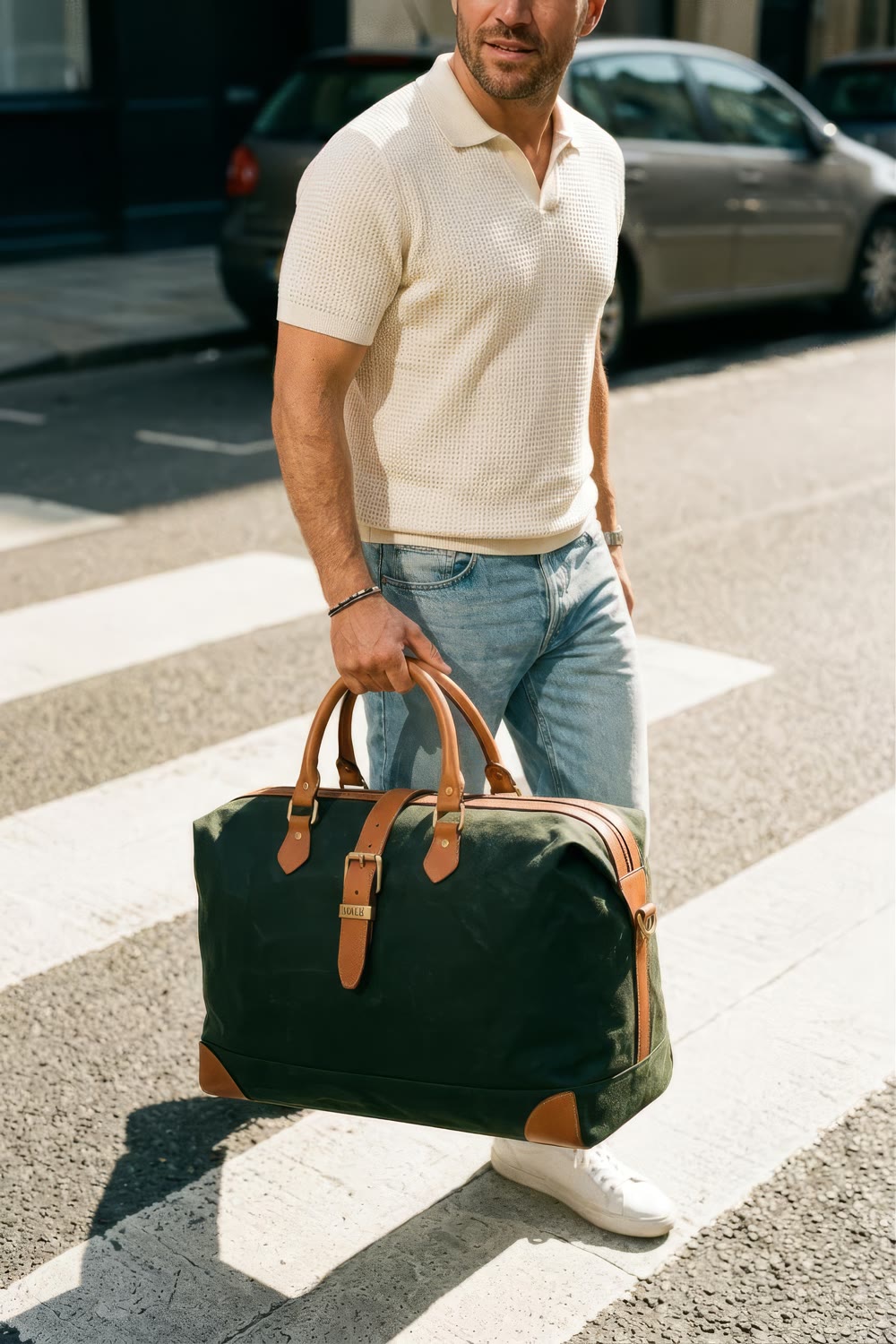 A man in a cream knit polo and jeans carrying the holdall across a zebra crossing