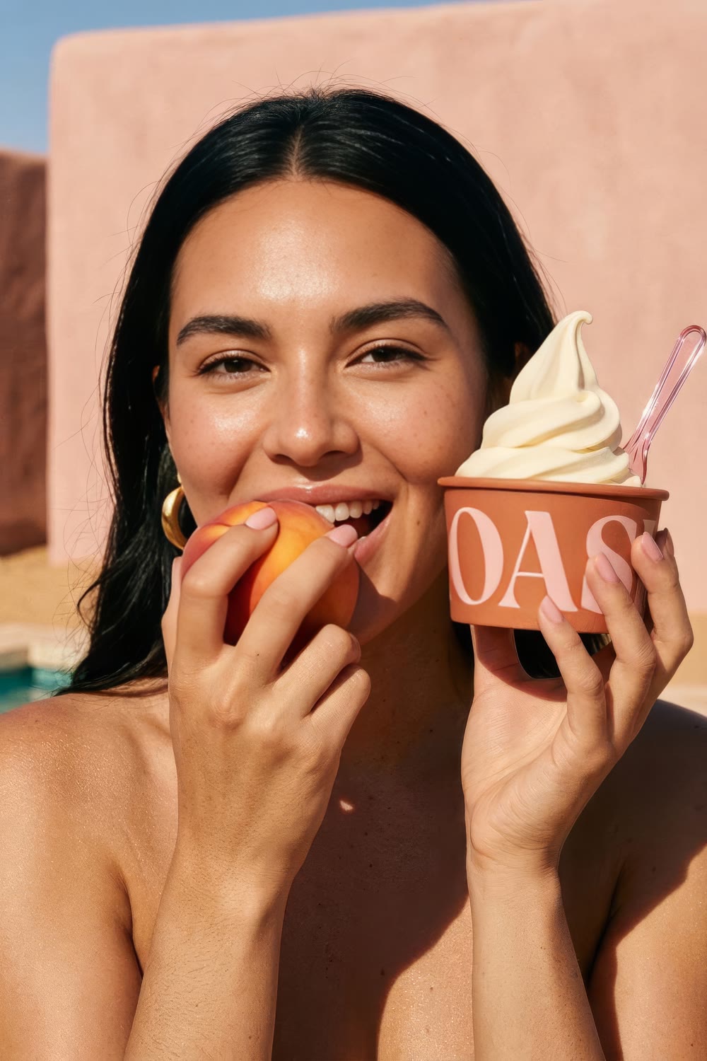 A woman biting a peach with the OASI cup held at her cheek against a pink adobe wall
