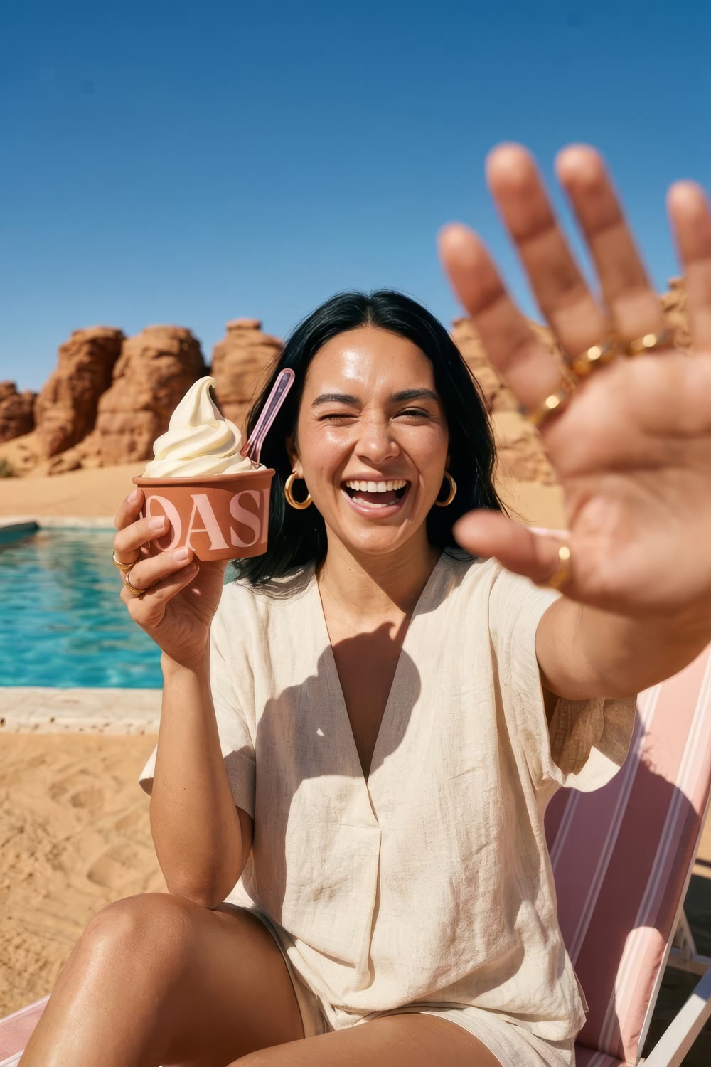A laughing woman on a striped lounger winking and holding the OASI cup, red desert rocks behind