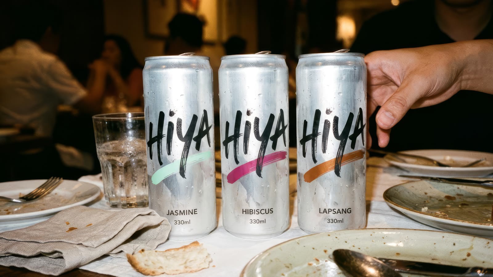 Three sparkling tea cans in jasmine, hibiscus and lapsang on a late dinner table with half-eaten plates, a hand reaching in