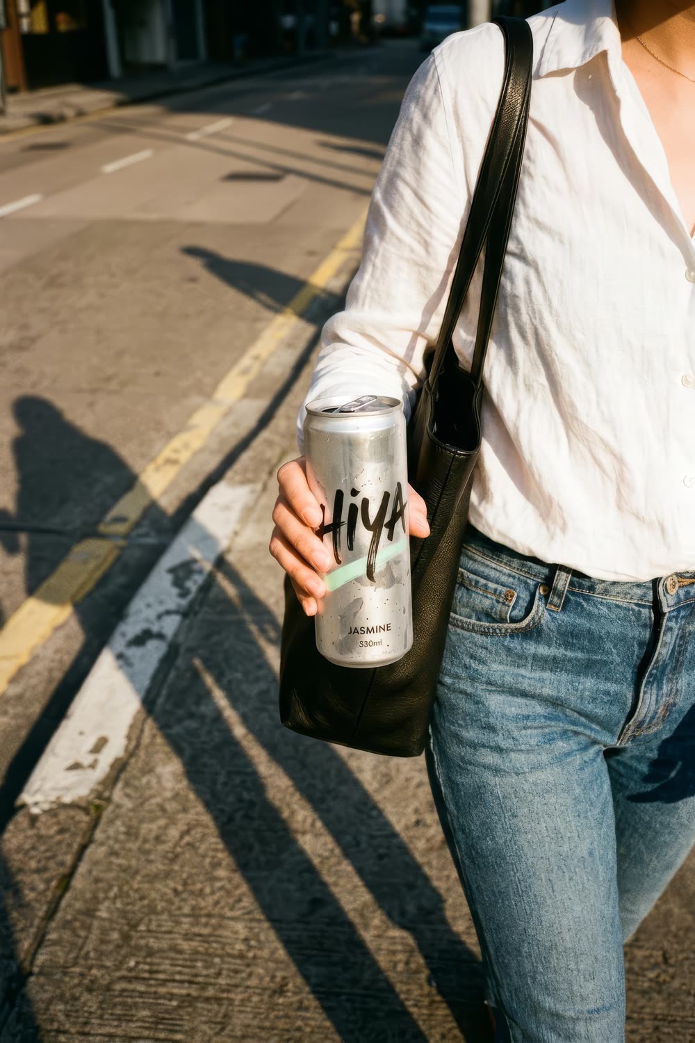 A woman in a white linen shirt and jeans walking with a leather tote, holding a jasmine can
