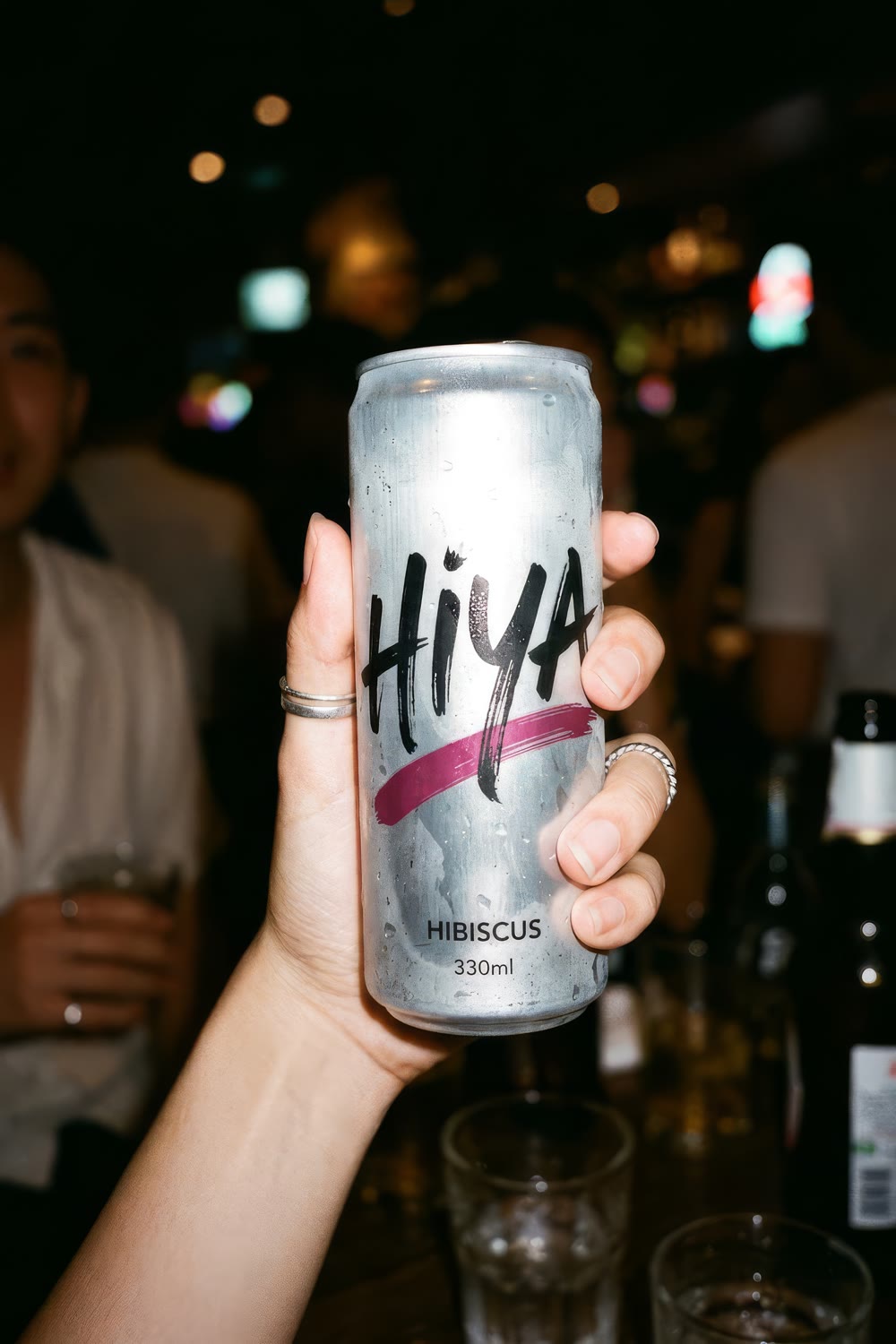 A hand with silver rings holding up a hibiscus can at a crowded bar in flash