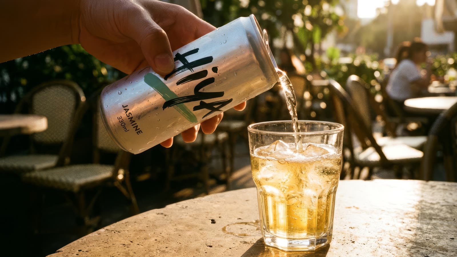 A hand pouring sparkling jasmine tea from the can into a glass of ice on a sunlit cafe table