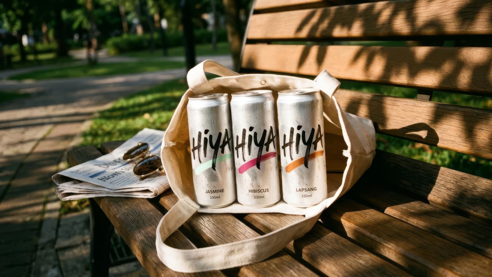 Three cans in a canvas tote on a sunlit park bench beside a newspaper and sunglasses