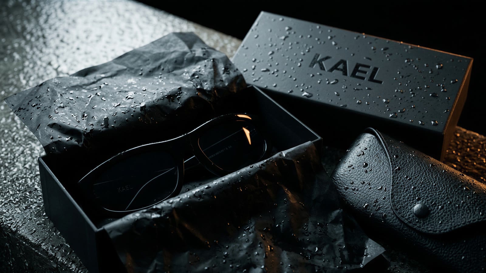 Sunglasses in an open matte black box with black tissue, a leather case and a KAEL-debossed lid
